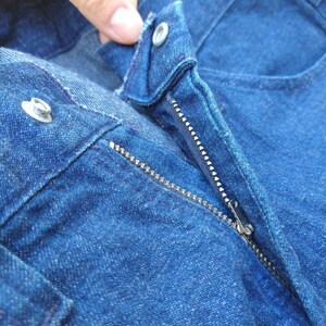 May include: Close-up of blue denim jeans with a silver zipper and button closure. The jeans are a medium wash and show signs of wear. The zipper is partially open, revealing the teeth.