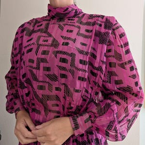 May include: A magenta dress with a high neckline and long sleeves. The dress features a geometric pattern in black and magenta. The fabric appears sheer, with a slight sheen. The dress has a cinched waist and a flowing skirt.