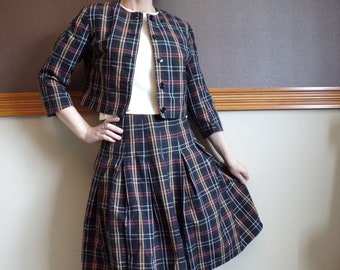 May include: A vintage plaid skirt suit featuring a cropped jacket and a pleated skirt. The suit has a dark background with a plaid pattern in red, tan, and white. The jacket has three buttons and three-quarter sleeves. The skirt falls to the knees.