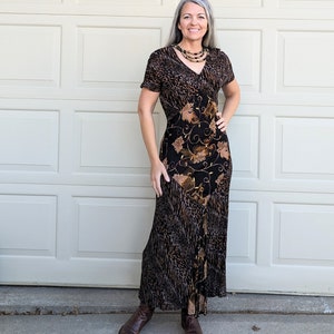May include: A long, flowing dress with a V-neck and short sleeves. The dress features a mix of patterns, including a leopard print and a floral design. The color palette includes brown, black, and gold tones. The model is wearing a multi-strand necklace and brown boots.