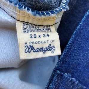 May include: Close-up of a Wrangler jeans tag. The tag is cream-colored with navy blue text that reads "29 x 34" and "A PRODUCT OF Wrangler." The tag also includes washing instructions. The jeans are blue denim.
