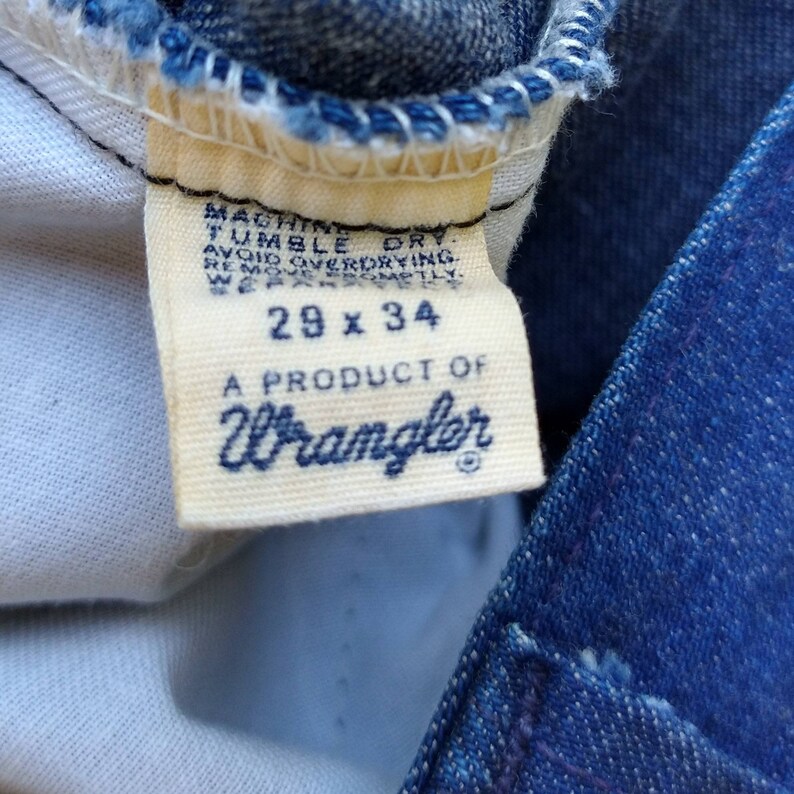 May include: Close-up of a Wrangler jeans tag. The tag is cream-colored with navy blue text that reads "29 x 34" and "A PRODUCT OF Wrangler." The tag also includes washing instructions. The jeans are blue denim.
