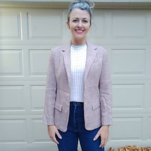 May include: A woman wearing a pink and white patterned blazer over a white sweater and blue jeans.