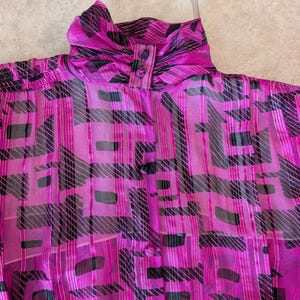 May include: A vibrant magenta blouse with a high, gathered collar and button closure. The sheer fabric features a bold geometric pattern in black and silver, creating a striking visual contrast. The blouse is a vintage piece, perfect for adding a pop of color and pattern to any outfit.