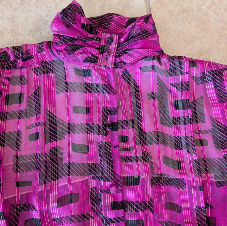 May include: A vibrant magenta blouse with a high, gathered collar and button closure. The sheer fabric features a bold geometric pattern in black and silver, creating a striking visual contrast. The blouse is a vintage piece, perfect for adding a pop of color and pattern to any outfit.