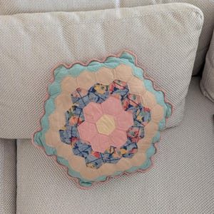 May include: A hexagonal patchwork pillow featuring a central yellow hexagon surrounded by pink, blue floral, and beige fabric pieces. The pillow is finished with a scalloped edge in light blue fabric, creating a layered, geometric quilted design.
