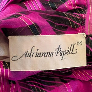 May include: Close-up of a vibrant pink fabric with black and gold accents. The fabric features a patterned design and the label reads "Adrianna Papell". The label is rectangular and has a cream background.