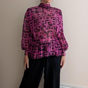 May include: A sheer, magenta blouse with a high neckline and long sleeves. The blouse features a black geometric pattern and a cinched waist. The model is wearing black pants.