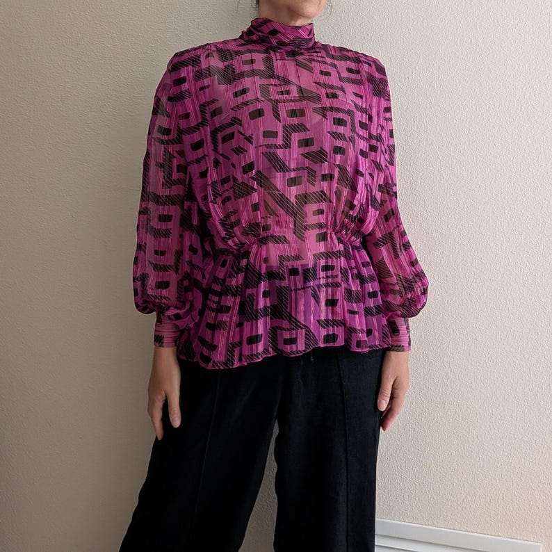 May include: A sheer, magenta blouse with a high neckline and long sleeves. The blouse features a black geometric pattern and a cinched waist. The model is wearing black pants.