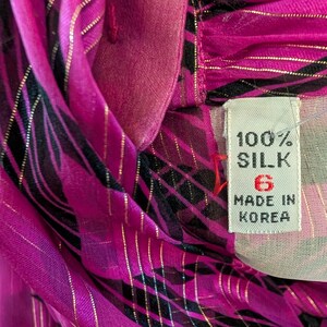 May include: Close-up of a vibrant pink silk garment with black and gold accents. The fabric is draped, revealing a tag that reads "100% SILK" and "MADE IN KOREA". The garment has a size 6.
