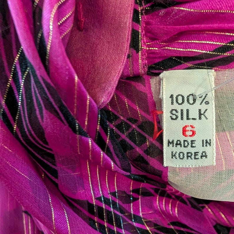 May include: Close-up of a vibrant pink silk garment with black and gold accents. The fabric is draped, revealing a tag that reads "100% SILK" and "MADE IN KOREA". The garment has a size 6.