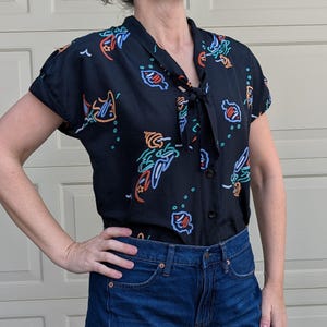 May include: Black short-sleeved blouse with a tie-neck collar and a button-down front. The blouse features a colorful neon-style print of sea creatures and other shapes. The model is wearing blue jeans.