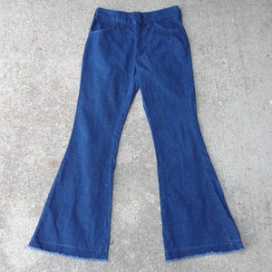 May include: Dark blue denim bell-bottom pants. The pants have a classic five-pocket design with a button and zipper closure. The flared legs have a raw hem. The pants are made of a medium-weight denim fabric.