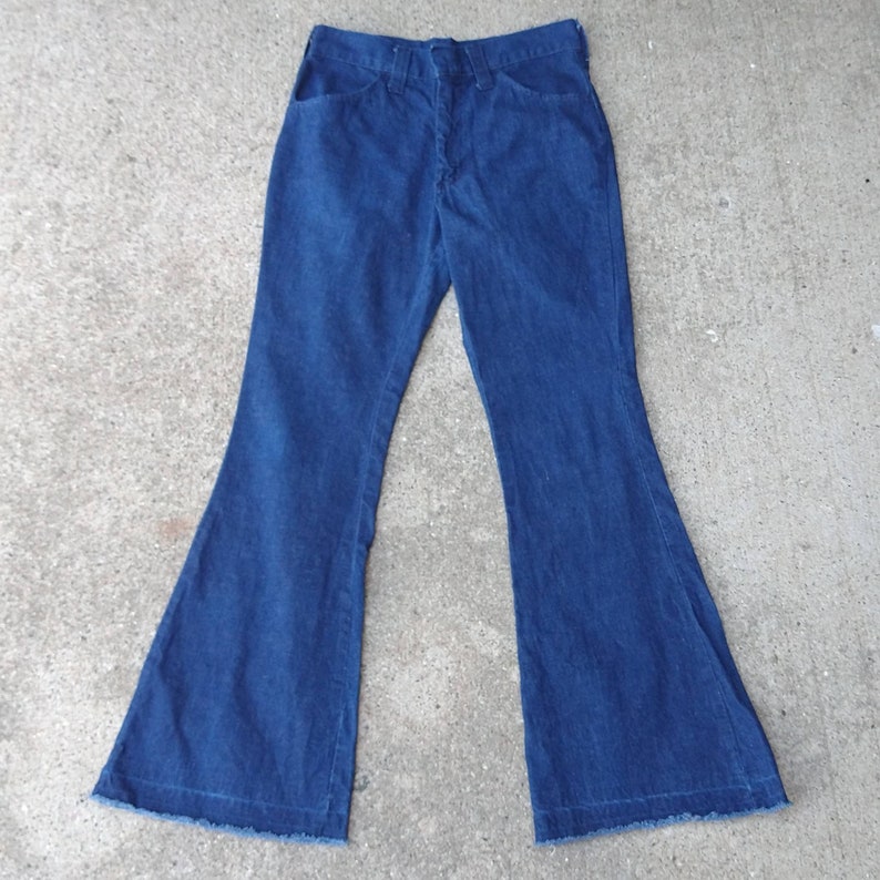 May include: Dark blue denim bell-bottom pants. The pants have a classic five-pocket design with a button and zipper closure. The flared legs have a raw hem. The pants are made of a medium-weight denim fabric.