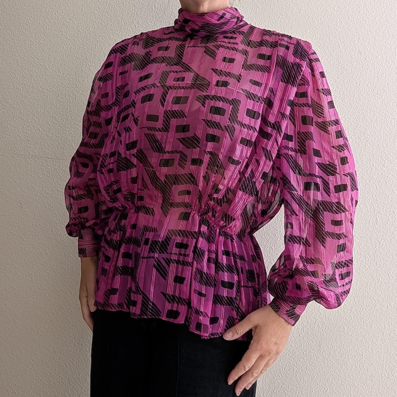 May include: A magenta blouse with a high neckline and long, voluminous sleeves. The sheer fabric features a geometric black pattern. The waist is cinched with elastic, creating a peplum effect. This vintage top is a stylish addition to any wardrobe.