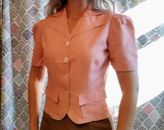 May include: A light pink short-sleeved blazer with a notched collar, two front pockets, and three white buttons. The blazer has a tailored fit and is made of a smooth fabric. The garment is part of a vintage clothing collection.