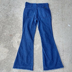 May include: Dark blue denim bell-bottom jeans. The jeans have a high waist, two back pockets, and a frayed hem. The Wrangler brand label is visible on the back pocket. The jeans are displayed on a concrete surface.