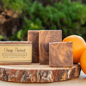 May include: Three rectangular bars of brown and tan marbled soap with a smooth texture. One bar features a beige paper label with the text Orange Patchouli Handcrafted Aloe Vera Soap Net Wt. Approx 5.5 oz / 156 g. Two whole oranges sit nearby.