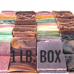 May include: A collection of handmade soap bars in various colors and textures. The bars are arranged in a box, with the text "1 LB. BOX" displayed prominently. The soaps feature shades of pink, green, brown, and purple, with visible layers and patterns.
