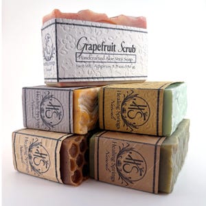 May include: A stack of five rectangular bars of soap wrapped in paper labels. The top label reads Grapefruit Scrub Handcrafted Aloe Vera Soap Net Wt. Approx 5.5 oz/156 g. The other labels feature the Healing Springs Naturals logo and text.