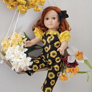 May include: A doll with red hair and a black bow sits on a wooden swing decorated with yellow and white flowers. The doll wears a yellow top and black overalls patterned with yellow sunflowers.