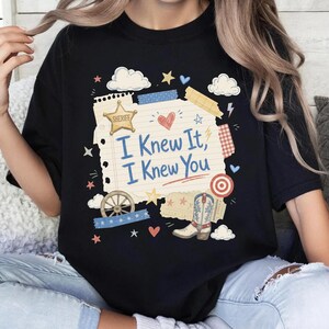 Op de afbeelding: Een zwart t-shirt met een afbeelding van een gelinieerd notitieblokvel met de tekst "I Knew It, I Knew You" in het blauw. Het ontwerp bevat een gouden sheriffster, een rood hart, een wagenwiel, een cowboylaars, wolken, sterren en een rood doelwit.