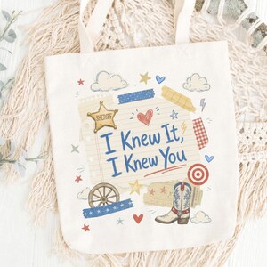 Op de afbeelding: Een cr&egrave;mekleurige canvas tas met een centrale afbeelding op een achtergrond van gelinieerd papier met de tekst "I Knew It, I Knew You". Het ontwerp bevat een gouden sheriffster, een cowboylaars, een wagenwiel, hartjes, sterren en wolken.