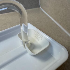 May include: A white plastic dish rack with a clear, curved handle. The rack has a small, attached compartment with the words "GLUTEN FREE" embossed on it. The rack is designed to hold dishes and is placed on a countertop.