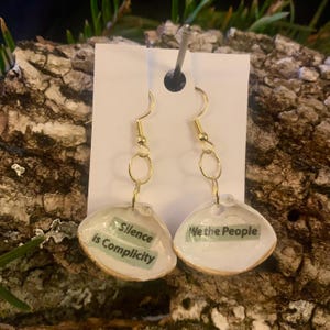 May include: A pair of seashell earrings with gold-colored hooks and connecting rings. Each shell has a printed message: "Silence is Complicity" and "We the People". The shells have a white interior and a gold-colored edge.