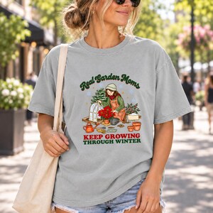 Op de afbeelding: Een lichtgrijs t-shirt met korte mouwen en een afbeelding van een persoon die tuiniert met een kerstster, een kleine kas en tuingereedschap. De tekst luidt Real Garden Moms KEEP GROWING THROUGH WINTER in donkergroene en rode letters.