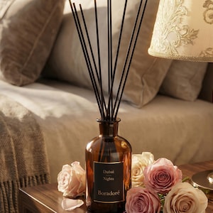 Pode incluir: Um frasco difusor de vidro castanho com varetas pretas, rotulado "Dubai Nights Boradoré", está sobre uma superfície de madeira com rosas cor-de-rosa e creme. Uma lâmpada decorativa com abajur estampado está no fundo.