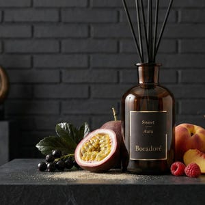 Puede incluir: Un difusor de vidrio marrón con varillas negras, etiquetado como "Sweet Aura Boradoré", se encuentra entre maracuyás, melocotones y frambuesas. El fondo oscuro resalta la botella ámbar y la fruta fresca, creando una estética lujosa.