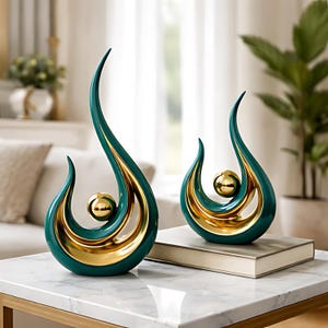 May include: Two abstract sculptures. Each has a teal exterior with gold-colored interior accents and a gold sphere. The sculptures have a teardrop shape with a curved, elongated top. They sit on a white marble surface.