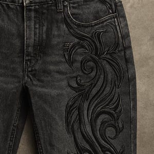 May include: Close-up of black denim jeans with an intricate, embossed floral design on the right leg. The jeans have a classic five-pocket design with a button and zipper closure. The dark wash denim has a slightly faded appearance.