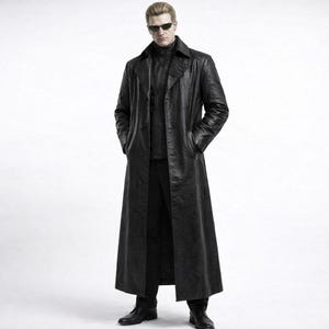 Op de afbeelding: Een lange, zwarte leren jas met een getextureerde afwerking. De jas heeft een lange, knoopsluiting, een reverskraag en zakken. De persoon op de afbeelding draagt een zwarte broek, een zwart overhemd en zwarte schoenen. De persoon draagt ook een zonnebril.