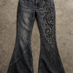 May include: Dark gray denim bell-bottom jeans with a faded wash. The jeans feature a decorative black embroidered design on the side of the leg. The jeans have a classic five-pocket design.