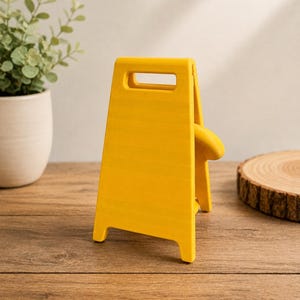 May include: A bright yellow, A-frame sign stand with a rectangular cutout handle at the top. The sign is constructed from a smooth, solid material and features a simple, upright design with four short legs, presented in a clean, minimalist aesthetic.