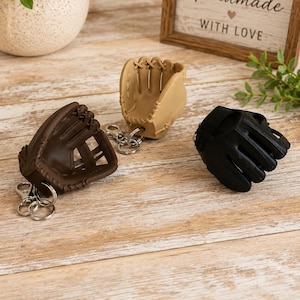 May include: Three baseball glove keychains in dark brown, tan, and black. Each keychain features a miniature leather-like glove with detailed stitching and a silver-toned metal lobster clasp attached to a circular split ring for keys.