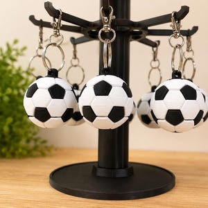 May include: A display of soccer ball keychains. Each keychain features a small, spherical soccer ball with black and white panels. The keychains are attached to a black display stand. The stand is on a wooden surface.