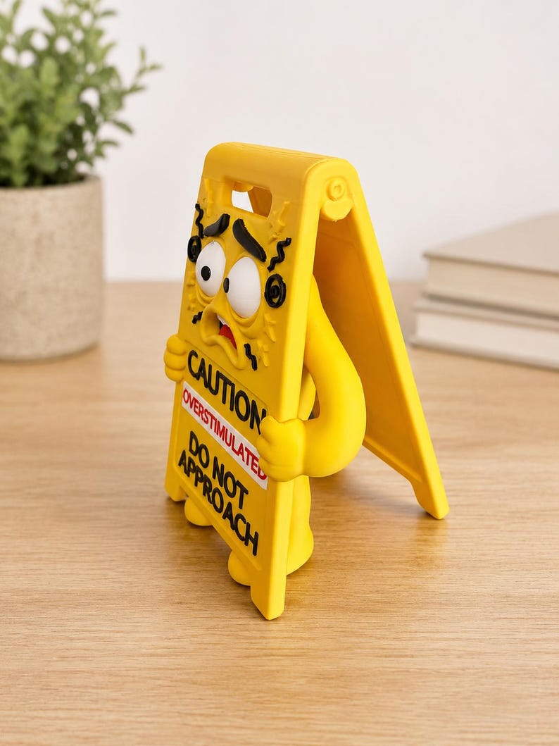 May include: A yellow A-frame caution sign figure with a distressed facial expression, featuring black text that reads CAUTION, OVERSTIMULATED, and DO NOT APPROACH. The figure has wide eyes, a small mouth, and arms holding the sign frame.