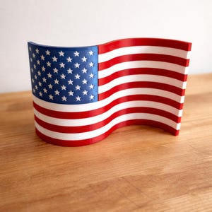May include: A curved, three-dimensional representation of the American flag featuring a blue canton with white stars on the left and alternating horizontal red and white stripes extending across the wavy, rigid structure of the object.