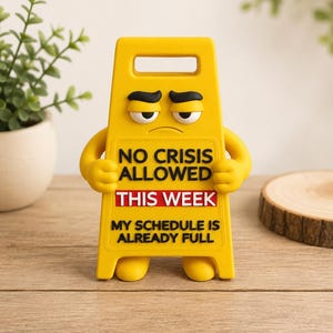 May include: A yellow plastic sign shaped like a caution floor sign with a cartoon face featuring furrowed brows. The text reads NO CRISIS ALLOWED THIS WEEK, with THIS WEEK in a red box, and MY SCHEDULE IS ALREADY FULL printed in black lettering.