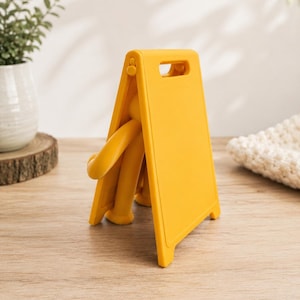 May include: A bright yellow plastic A-frame sign with a smooth, matte finish. The sign features a rectangular panel with a cutout handle at the top and a hinged design that allows it to stand upright. A small, yellow, articulated figure is visible.