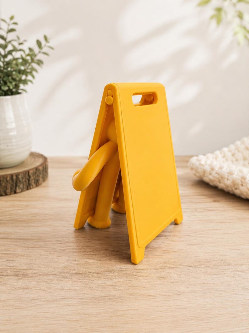 May include: A bright yellow plastic A-frame sign with a smooth, matte finish. The sign features a rectangular panel with a cutout handle at the top and a hinged design that allows it to stand upright. A small, yellow, articulated figure is visible.