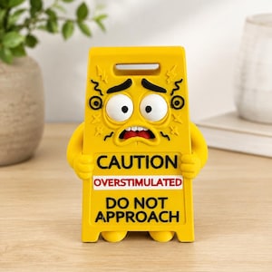 May include: A yellow plastic caution sign shaped like a character with a stressed facial expression, large white eyes, and black eyebrows. The text reads CAUTION OVERSTIMULATED DO NOT APPROACH in black and red lettering on the front of the sign.