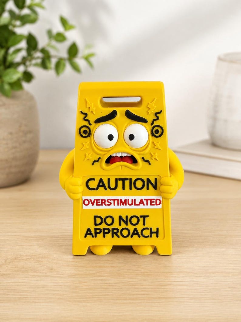 May include: A yellow plastic caution sign shaped like a character with a stressed facial expression, large white eyes, and black eyebrows. The text reads CAUTION OVERSTIMULATED DO NOT APPROACH in black and red lettering on the front of the sign.