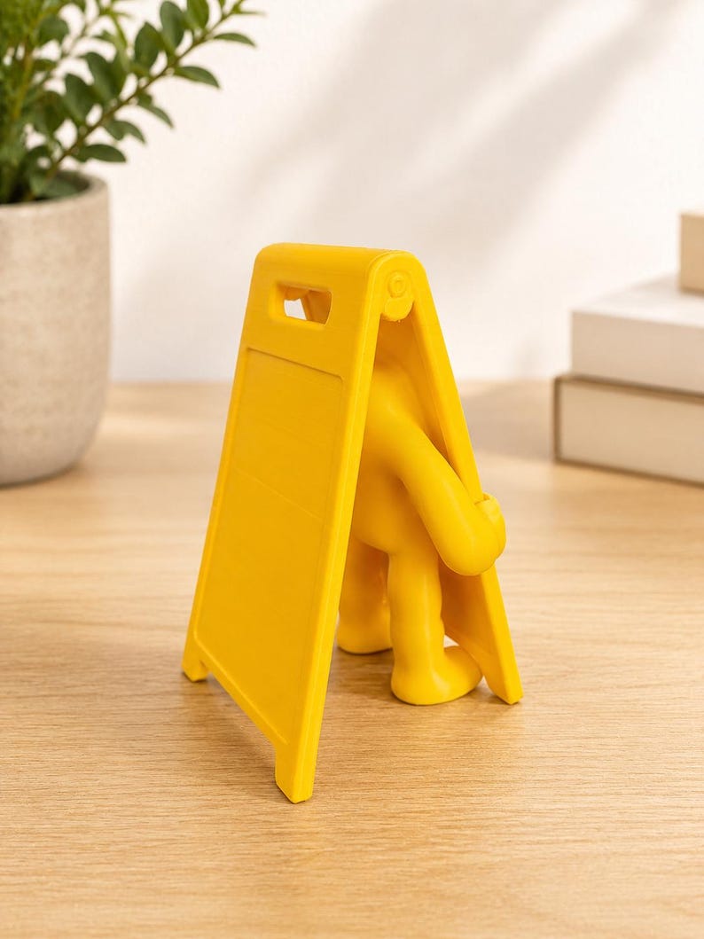 May include: A bright yellow plastic figure shaped like a person holding a blank A-frame sign. The figure has a smooth, matte finish and stands in a tripod position, with the sign panel angled forward and a handle cutout at the top of the frame.