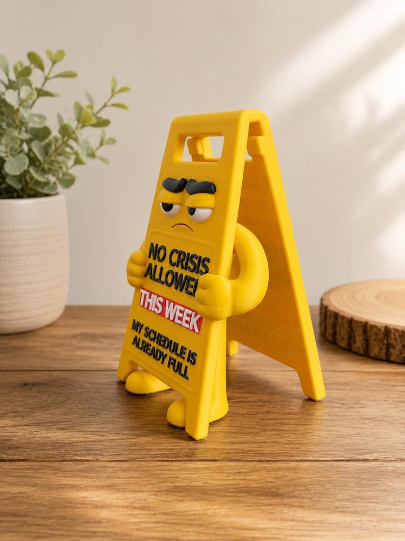 May include: A yellow A-frame sign figurine with a grumpy facial expression, featuring the black text NO CRISIS ALLOWED, the red and white text THIS WEEK, and the black text MY SCHEDULE IS ALREADY FULL printed on the front surface.