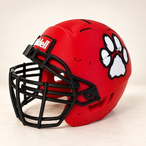 May include: A matte red football helmet featuring a black face mask and a white paw print decal on the side. The Riddell brand name is printed in white on a red rectangular background located above the face mask on the front of the helmet.