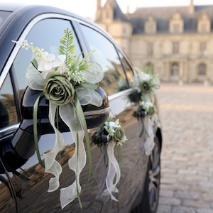 Op de afbeelding: Een decoratief bloemstuk bevestigd aan de zijspiegel van een zwarte auto, met een grote saliegroene roos, witte bloesems, groene varenbladeren en lange, zwierige linten van doorschijnende witte en saliegroene stof in een watervalvorm.